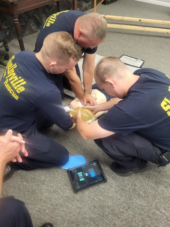 Child CPR training: rescuers check airway on mannequin with smartphone timer on mat.