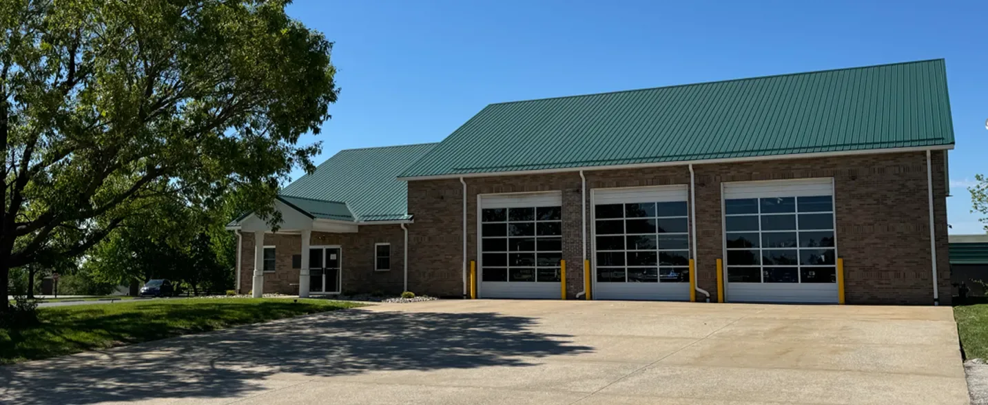 Modern institutional building with green metal roof, large windows, and sunny parking lot
